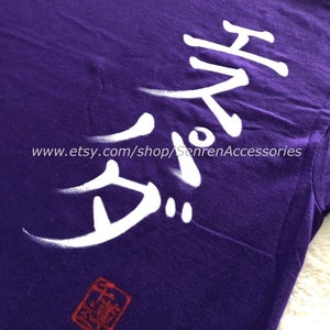 May include: A purple t-shirt with white Japanese characters and a red stamp. The text is stylized and appears to be hand-painted. The Etsy shop URL is visible at the bottom of the image.