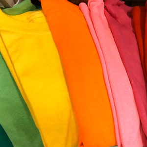 May include: A collection of colorful t-shirts in shades of green, yellow, orange, pink, and red. The shirts are neatly folded, showcasing their vibrant hues and soft fabric. The image highlights the variety of colors available.