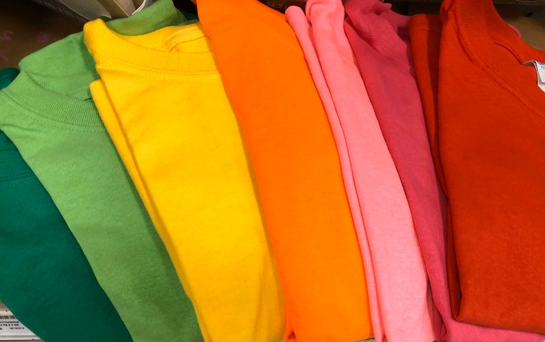 May include: A collection of colorful t-shirts in shades of green, yellow, orange, pink, and red. The shirts are neatly folded, showcasing their vibrant hues and soft fabric. The image highlights the variety of colors available.