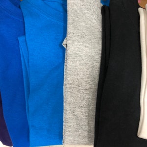May include: A collection of folded t-shirts in various colors, including purple, blue, teal, gray, black, and white. The shirts are neatly stacked, showcasing a range of hues and styles. The image highlights the variety of available colors.