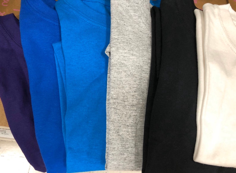 May include: A collection of folded t-shirts in various colors, including purple, blue, teal, gray, black, and white. The shirts are neatly stacked, showcasing a range of hues and styles. The image highlights the variety of available colors.