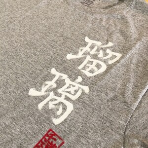 May include: A heather gray Gildan t-shirt featuring white Japanese characters and a red square stamp. The shirt has a crew neck and short sleeves. The Japanese characters are vertically aligned.