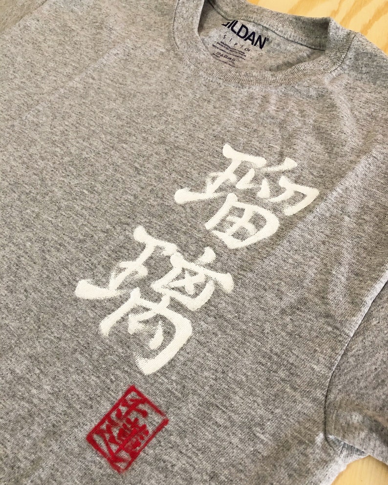 May include: A heather gray Gildan t-shirt featuring white Japanese characters and a red square stamp. The shirt has a crew neck and short sleeves. The Japanese characters are vertically aligned.