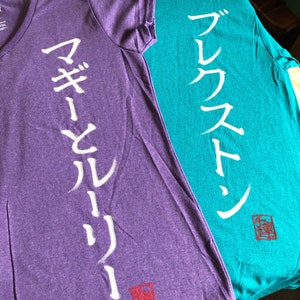 May include: Two t-shirts, one purple and one teal, are displayed. The purple shirt has a V-neck and white Japanese characters down the front. The teal shirt has a crew neck and white Japanese characters. Both shirts have a red square symbol.