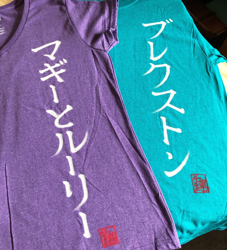 May include: Two t-shirts, one purple and one teal, are displayed. The purple shirt has a V-neck and white Japanese characters down the front. The teal shirt has a crew neck and white Japanese characters. Both shirts have a red square symbol.