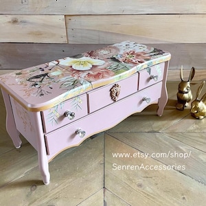 May include: A pink jewelry box with three drawers and a floral design on the top. The box has crystal knobs and gold accents. Two gold rabbit figurines are next to the box. The text "www.etsy.com/shop/SenrenAccessories" is visible.
