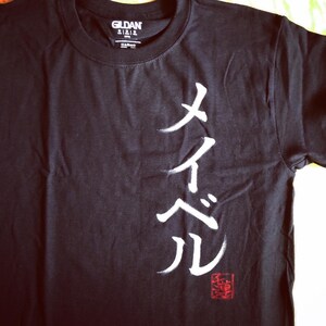 May include: Black Gildan t-shirt with white Japanese calligraphy and a red stamp. The shirt features a crew neck and short sleeves. The Japanese characters are the focal point of the design.