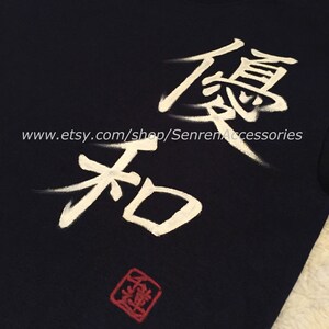 May include: Navy blue t-shirt with white Japanese calligraphy and a red square stamp. The text includes the characters for "excellence" and "harmony". The Etsy shop URL is visible at the bottom of the shirt.