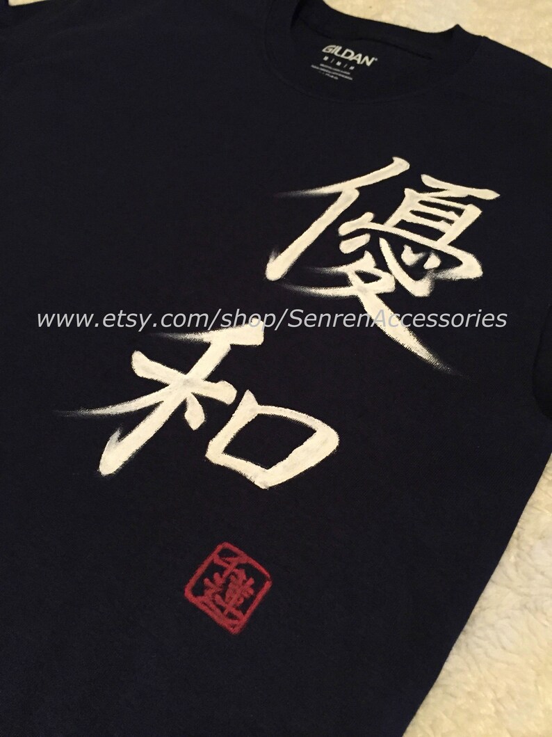 May include: Navy blue t-shirt with white Japanese calligraphy and a red square stamp. The text includes the characters for "excellence" and "harmony". The Etsy shop URL is visible at the bottom of the shirt.