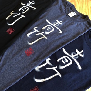 May include: Three t-shirts in black, heather gray, and navy blue, each featuring a white, stylized Japanese character design. A red square stamp is below the design on each shirt. The shirts are made of a soft material.