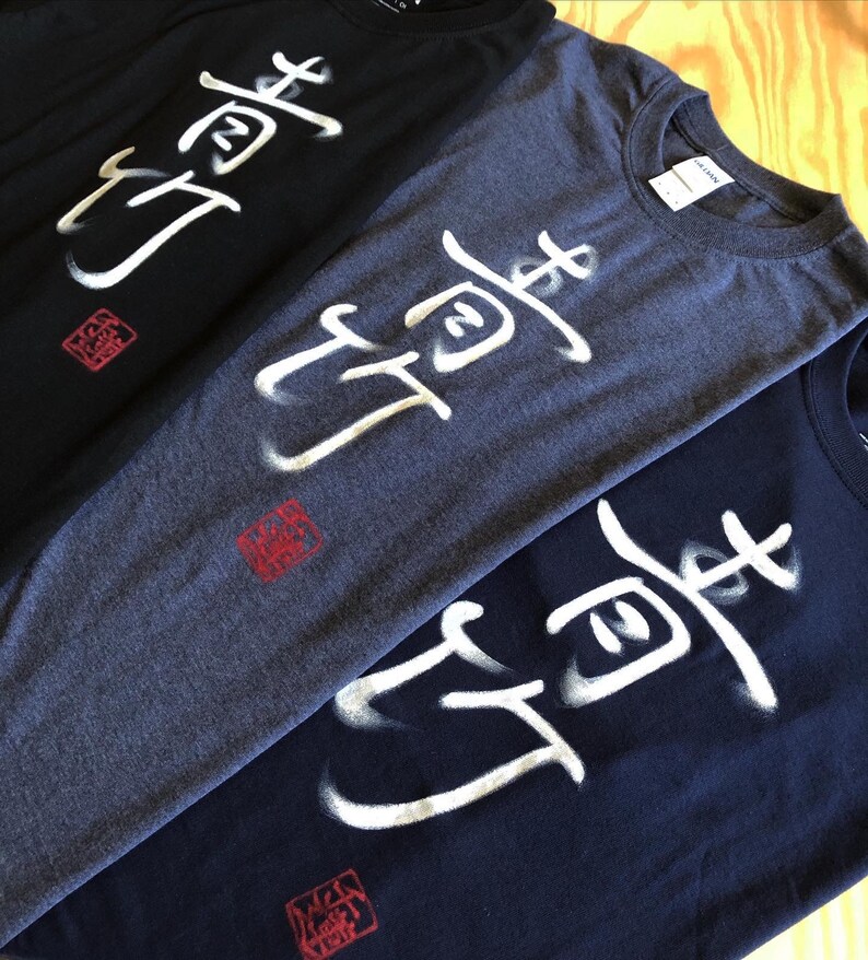 May include: Three t-shirts in black, heather gray, and navy blue, each featuring a white, stylized Japanese character design. A red square stamp is below the design on each shirt. The shirts are made of a soft material.