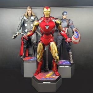 Op de afbeelding: Drie actiefiguren van Marvel-superhelden: Iron Man, Thor en Captain America. Iron Man, in een rood en goudkleurig pak, staat in het midden. Thor, met een hamer, staat links. Captain America, met een schild, staat rechts. Elke figuur staat op een zwarte basis.