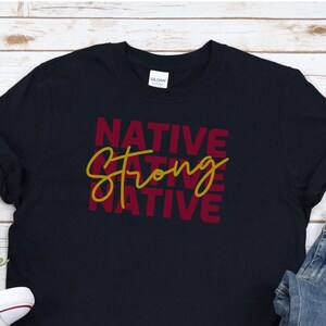 May include: A dark blue t-shirt with the words "NATIVE STRONG NATIVE" printed in maroon and gold. The shirt is folded and laid on a wooden surface with a pair of white sneakers and blue jeans.
