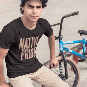 May include: Black t-shirt with the text "NATIVE PRIDE" in a distressed, vintage style. The shirt is worn by a person sitting next to a blue BMX bike. The text is in a beige and red color.