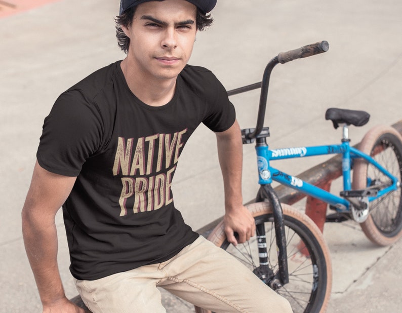 May include: Black t-shirt with the text "NATIVE PRIDE" in a distressed, vintage style. The shirt is worn by a person sitting next to a blue BMX bike. The text is in a beige and red color.