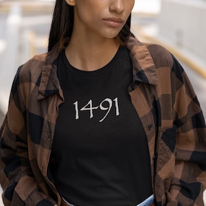 May include: A person wearing a black t-shirt with the number "1491" printed on it. They are also wearing a brown and black plaid shirt and blue jeans.