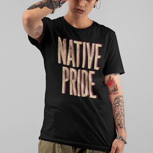 May include: Black t-shirt with the words "NATIVE PRIDE" in a distressed, vintage-style font. The text is in a cream color with a pink shadow. The shirt is short-sleeved and worn by a person with tattoos and piercings.