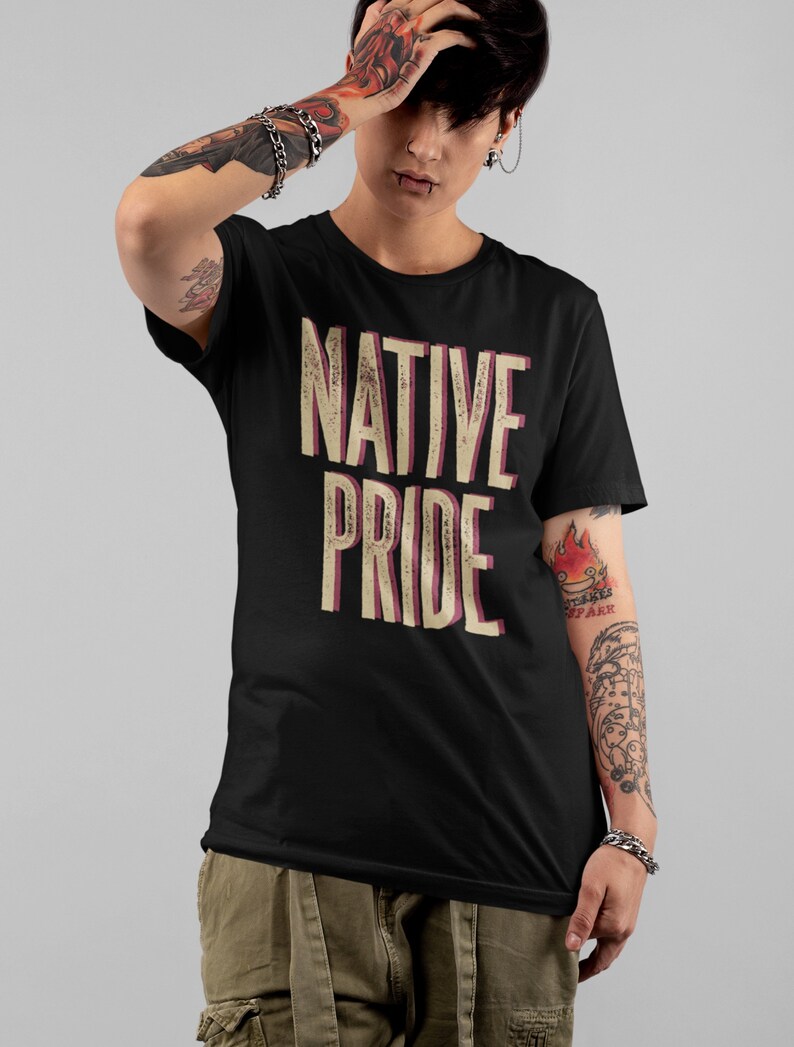 May include: Black t-shirt with the words "NATIVE PRIDE" in a distressed, vintage-style font. The text is in a cream color with a pink shadow. The shirt is short-sleeved and worn by a person with tattoos and piercings.