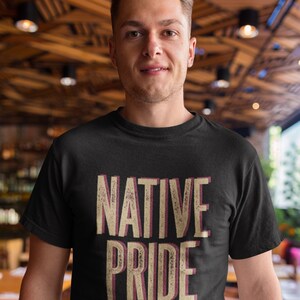 May include: Black crew neck t-shirt with the words "NATIVE PRIDE" in a distressed, layered font. The text is in beige with a maroon shadow. The shirt is worn by a person with short brown hair.