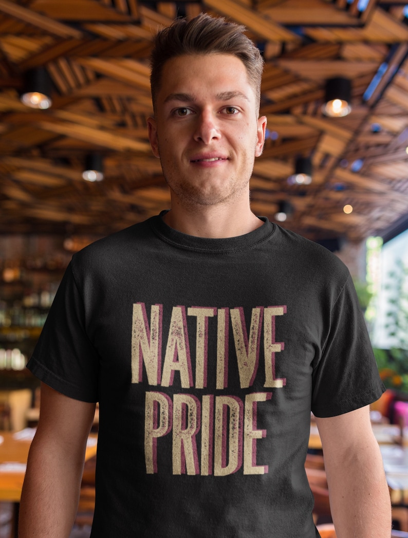May include: Black crew neck t-shirt with the words "NATIVE PRIDE" in a distressed, layered font. The text is in beige with a maroon shadow. The shirt is worn by a person with short brown hair.
