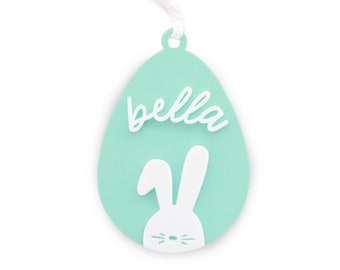 May include: A light blue Easter egg-shaped ornament with a white bunny and the word "bella" in white script.