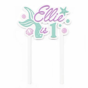 May include: A cake topper with a mermaid tail, a seashell, and a star. The topper says "Ellie is 1" in purple and green lettering.