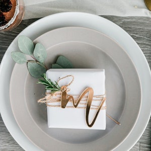 May include: A white gift box with a gold letter 'M' on top, tied with twine and a sprig of rosemary. The box is on a gray plate, which is on a white plate. The plates are on a wooden table with a green cloth napkin.