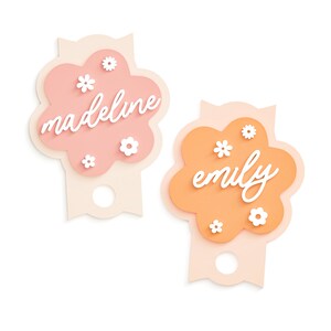 May include: Two pink and orange paper gift tags shaped like flowers with white paper flowers and the names "madeline" and "emily" written in white.