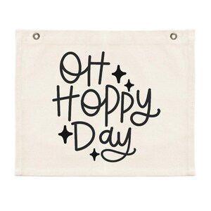 May include: A white canvas wall hanging with black text that reads "Oh Hoppy Day" with small star accents.