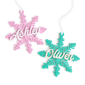 May include: Two snowflake ornaments with names "Ashley" and "Oliver" written on them. The snowflake on the left is pink and the snowflake on the right is teal.