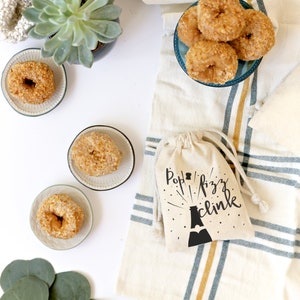Pu&ograve; includere: Una borsa di stoffa bianca con il testo "Pop Fizz Clink" e una silhouette nera di una bottiglia di champagne con un design a stella. La borsa &egrave; appoggiata su un tessuto a strisce bianche e blu. Ci sono anche dei donut su un piatto blu e una piccola pianta grassa in un vaso.