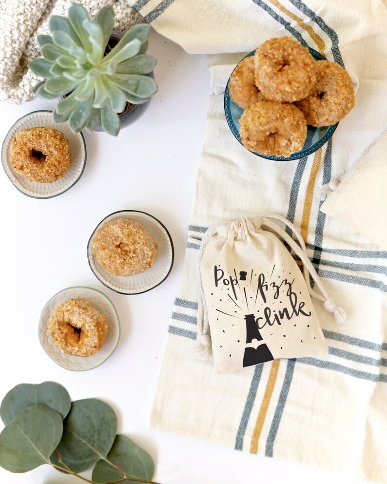 Pu&ograve; includere: Una borsa di stoffa bianca con il testo "Pop Fizz Clink" e una silhouette nera di una bottiglia di champagne con un design a stella. La borsa &egrave; appoggiata su un tessuto a strisce bianche e blu. Ci sono anche dei donut su un piatto blu e una piccola pianta grassa in un vaso.