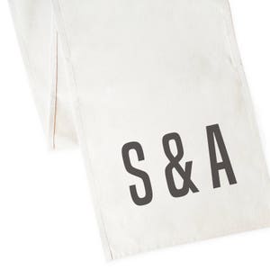May include: White linen table runner with the initials "S&A" printed in black.
