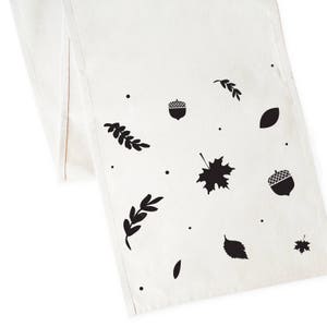 May include: White table runner with a black and white autumn leaf and acorn design. The design includes leaves, acorns, and branches.