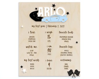 May include: A wooden sign with a blue and white car graphic and the name "ARLO" in black letters. The sign lists details about the child's first year, including their weight, height, favorite foods, toys, and words.