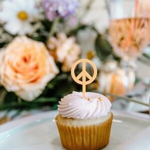 Pu&ograve; includere: Un cupcake con glassa rosa e un topper a forma di simbolo della pace. Il cupcake &egrave; su un piatto bianco, con fiori e una bevanda sullo sfondo. Il simbolo della pace &egrave; di colore marrone chiaro.