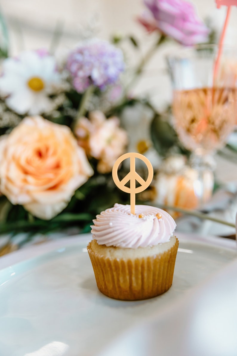 Pu&ograve; includere: Un cupcake con glassa rosa e un topper a forma di simbolo della pace. Il cupcake &egrave; su un piatto bianco, con fiori e una bevanda sullo sfondo. Il simbolo della pace &egrave; di colore marrone chiaro.