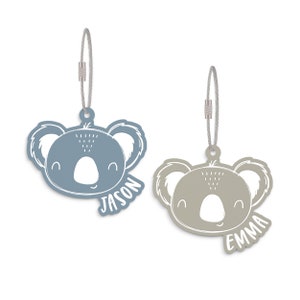 May include: Two koala-shaped luggage tags with silver metal rings. The tag on the left is blue with the name "JASON" written on it. The tag on the right is beige with the name "EMMA" written on it.