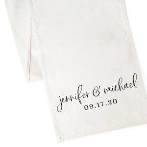 May include: White linen table runner with the text "jennifer & michael 09.17.20" printed in black.