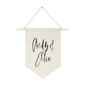 May include: A white canvas banner with a wooden dowel at the top and a black inscription that reads "Andy & Ellie" in a cursive font.