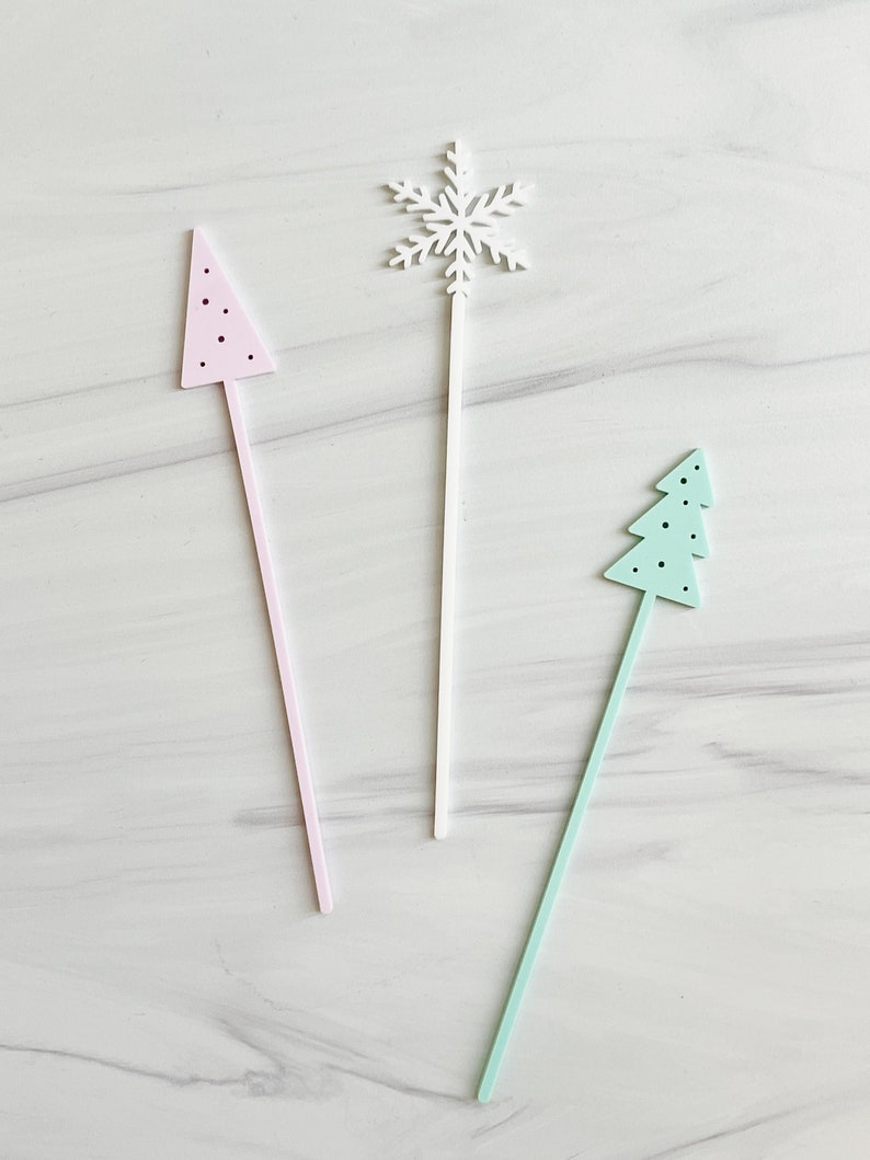 May include: Three wooden stick ornaments with different holiday shapes: a pink Christmas tree, a white snowflake, and a light blue Christmas tree. The ornaments are on a white marble background.