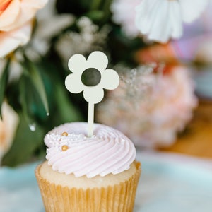 Pu&ograve; includere: Un cupcake con glassa rosa chiaro, guarnito con spruzzi dorati e una decorazione a forma di fiore. Il cupcake &egrave; su una superficie azzurra, con fiori sullo sfondo.