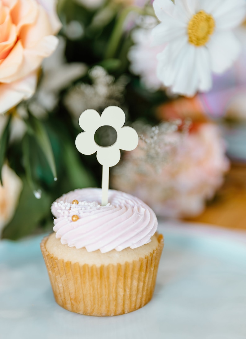 Pu&ograve; includere: Un cupcake con glassa rosa chiaro, guarnito con spruzzi dorati e una decorazione a forma di fiore. Il cupcake &egrave; su una superficie azzurra, con fiori sullo sfondo.