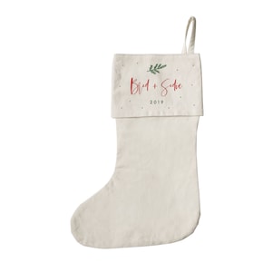 May include: A white Christmas stocking with a red and green design. The stocking has the names "Brad + Sadie" and the year "2019" printed on it.