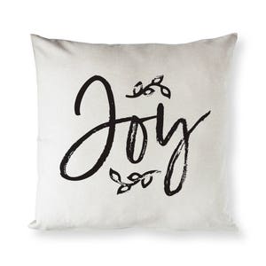May include: A white throw pillow with black brushstroke lettering that says "Joy" with a small floral design above and below the word.