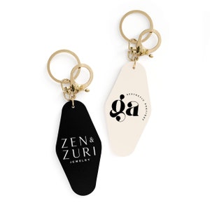 May include: Two black and white motel keychains with gold key rings. One keychain says "Zen & Zuri Jewelry" and the other says "ga Aesthetic Boutique".