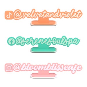 May include: Three social media display stands in orange, teal, and pink. Each stand features a different social media icon and handle: @velvetandviolet, @serenesoulspa, and @bloomblisscafe. The stands are designed to be freestanding.