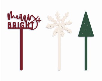 May include: Three festive cake toppers: a red "Merry & Bright" sign, a white snowflake, and a green Christmas tree.