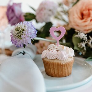 Pu&ograve; includere: Un cupcake con glassa rosa e un decoro a forma di cuore rosa &egrave; su un piatto bianco. Lo sfondo sfocato presenta fiori rosa, pesca e viola e un tovagliolo bianco.