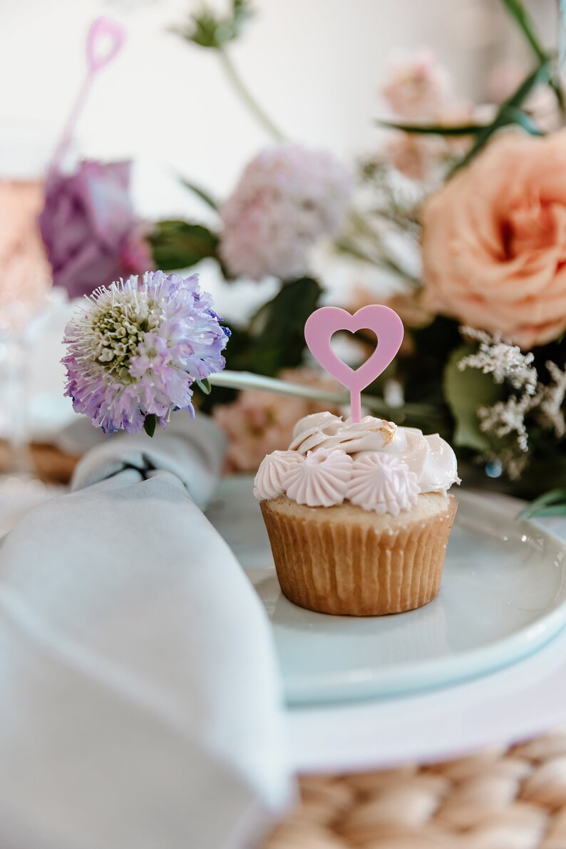 Pu&ograve; includere: Un cupcake con glassa rosa e un decoro a forma di cuore rosa &egrave; su un piatto bianco. Lo sfondo sfocato presenta fiori rosa, pesca e viola e un tovagliolo bianco.